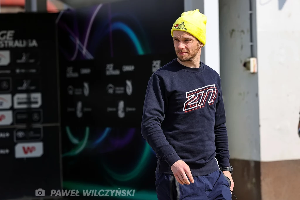 Krzysztof Buczkowski przed zawodami. Mężczyzna w żółtej czapce i granatowej bluzie z dużym nadrukiem stoi na tle ściany z logotypami sponsorów oraz kolorowymi światłami w tle.