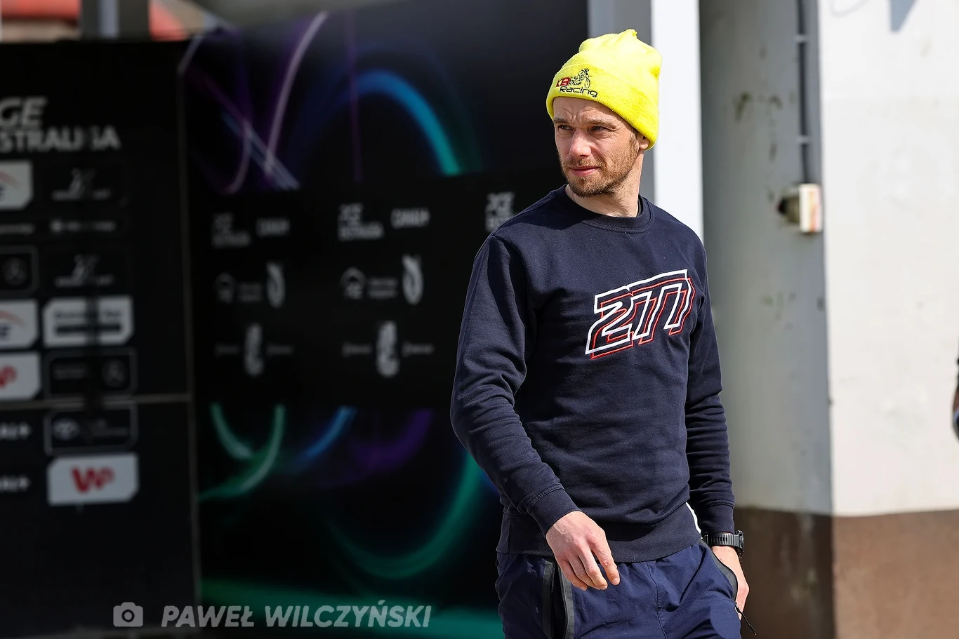 Mężczyzna w żółtej czapce i granatowej bluzie z dużym nadrukiem stoi na tle ściany z logotypami sponsorów oraz kolorowymi światłami w tle.
