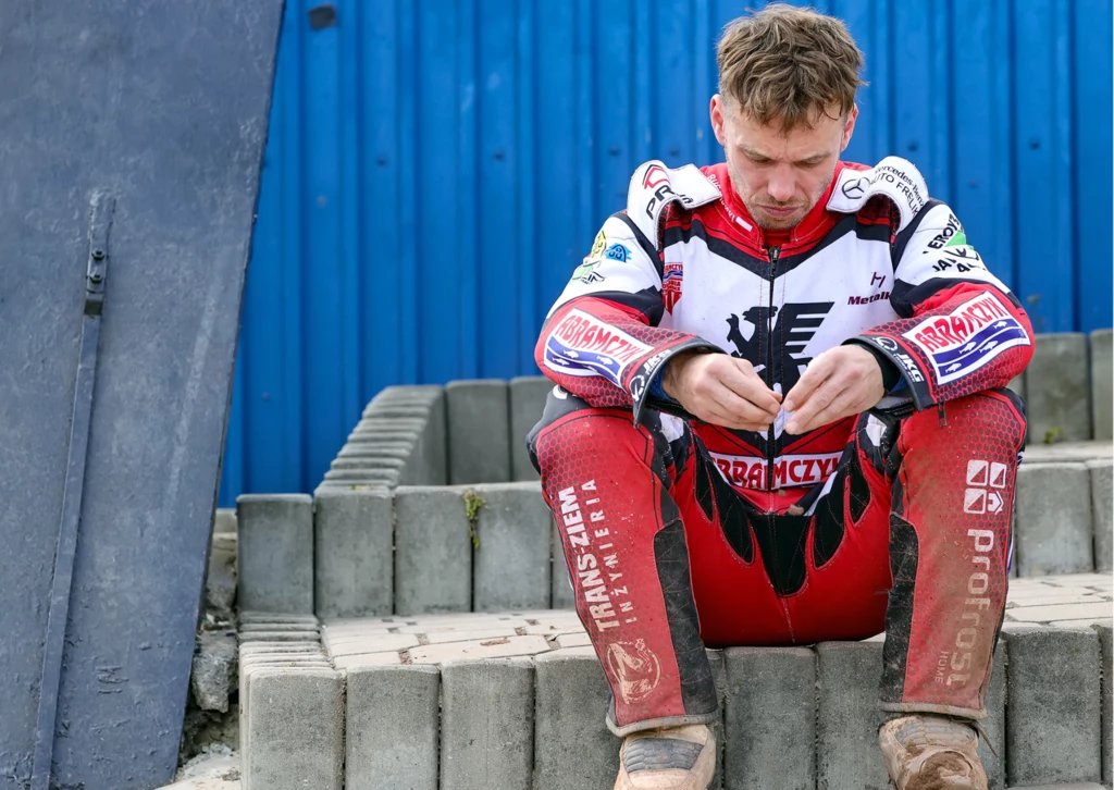 Krzysztof Buczkowski załamany złym występem w lidze. Zawodnik żużlowy w pełnym stroju sportowym siedzi na betonowych schodkach ze spuszczoną głową, sprawia wrażenie zamyślonego lub zamartwionego, w tle widoczna niebieska blacha.