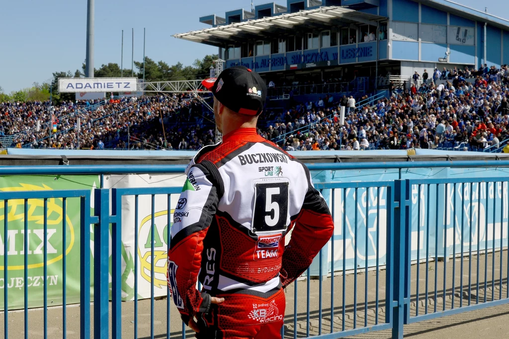 Krzysztof Buczkowski na stadionie w Lesznie. Zawodnik żużlowy w czerwonym kombinezonie z numerem 5 na plecach stoi przy niebieskim ogrodzeniu, patrząc w stronę trybun wypełnionych kibicami na stadionie sportowym. W tle widoczna nowoczesna trybuna z zadaszeniem.