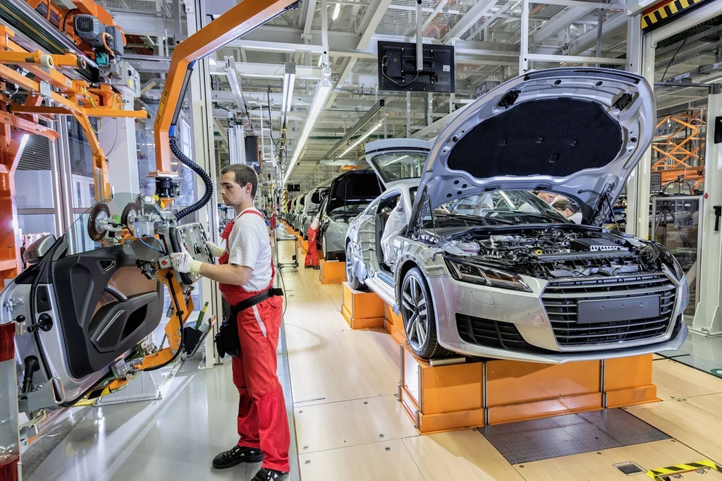 Audi to jedna z firm najbardziej dotkniętych nowymi cłami USA. Montaż samochodów na nowoczesnej linii produkcyjnej, pracownik w czerwonych spodniach obsługuje maszynę montującą drzwi, w tle widoczne kolejne pojazdy w różnych etapach składania.
