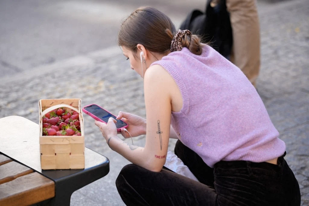 mObywatel - Ministerstwo Cyfryzacji tłumaczy prośbę o dostęp do biometrii, kamery i lokalizacji Młoda kobieta w liliowym swetrze siedzi na ławce, przeglądając telefon komórkowy, obok koszyk pełen świeżych truskawek