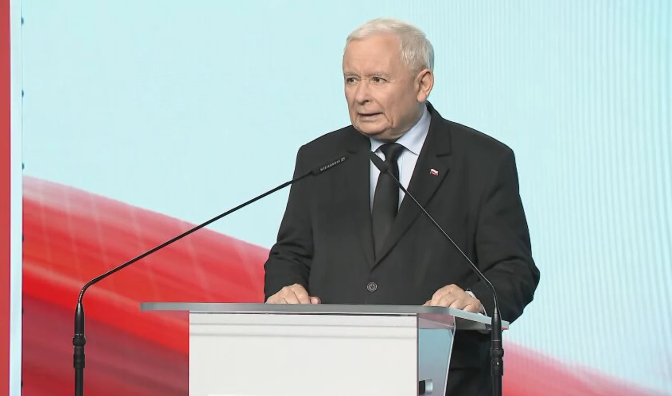 Prezes PiS Jarosław Kaczyński Starszy mężczyzna w czarnym garniturze i białej koszuli przemawia przy przezroczystej mównicy, na tle dużego ekranu z biało-czerwonym motywem.