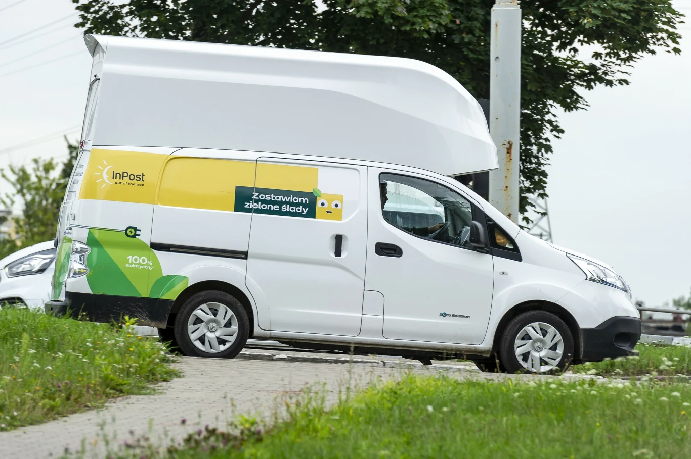 Zarzuty UOKiK dotyczą m.in. "zielonego" przekazu marketingowego firm kurierskich Samochód dostawczy firmy InPost z widocznym logo oraz hasłem reklamowym na boku pojazdu, zaparkowany na chodniku w pobliżu ulicy, w tle drzewa oraz fragment innego samochodu.