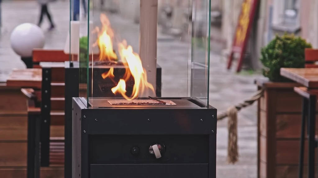 Nowoczesny ogrzewacz gazowy ogrodowy z widocznym ogniem, otoczony szklanymi panelami, ustawiony na tle drewnianych mebli ogrodowych i dekoracyjnych roślin przy restauracyjnym ogródku na miejskiej ulicy.