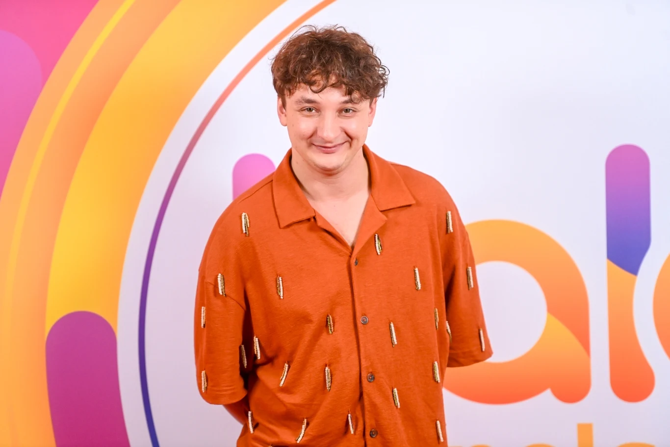 Wojtek Szumański prezentuje nowy teledysk "Jakoś to będzie" Mężczyzna o kręconych włosach ubrany w pomarańczową koszulę z wzorem, stojący na tle kolorowych elementów graficznych i dużego napisu