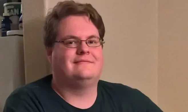 Jason Kingsford / JustInBasil Mężczyzna w okularach z krótkimi, jasnobrązowymi włosami uśmiecha się lekko, ubrany w ciemną koszulkę, siedzi w pomieszczeniu z jasną ścianą i widocznymi przedmiotami na półce w tle.