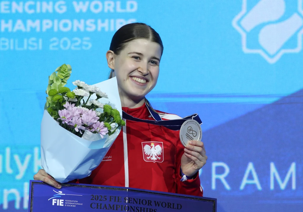 Zuzanna Cieślar ze srebrnym medalem mistrzostw świata Uśmiechnięta kobieta w czerwonym sportowym dresie z godłem Polski trzyma w jednej ręce bukiet kwiatów, a w drugiej medal, za nią znajduje się tło z nazwą i logiem mistrzostw świata w szermierce.