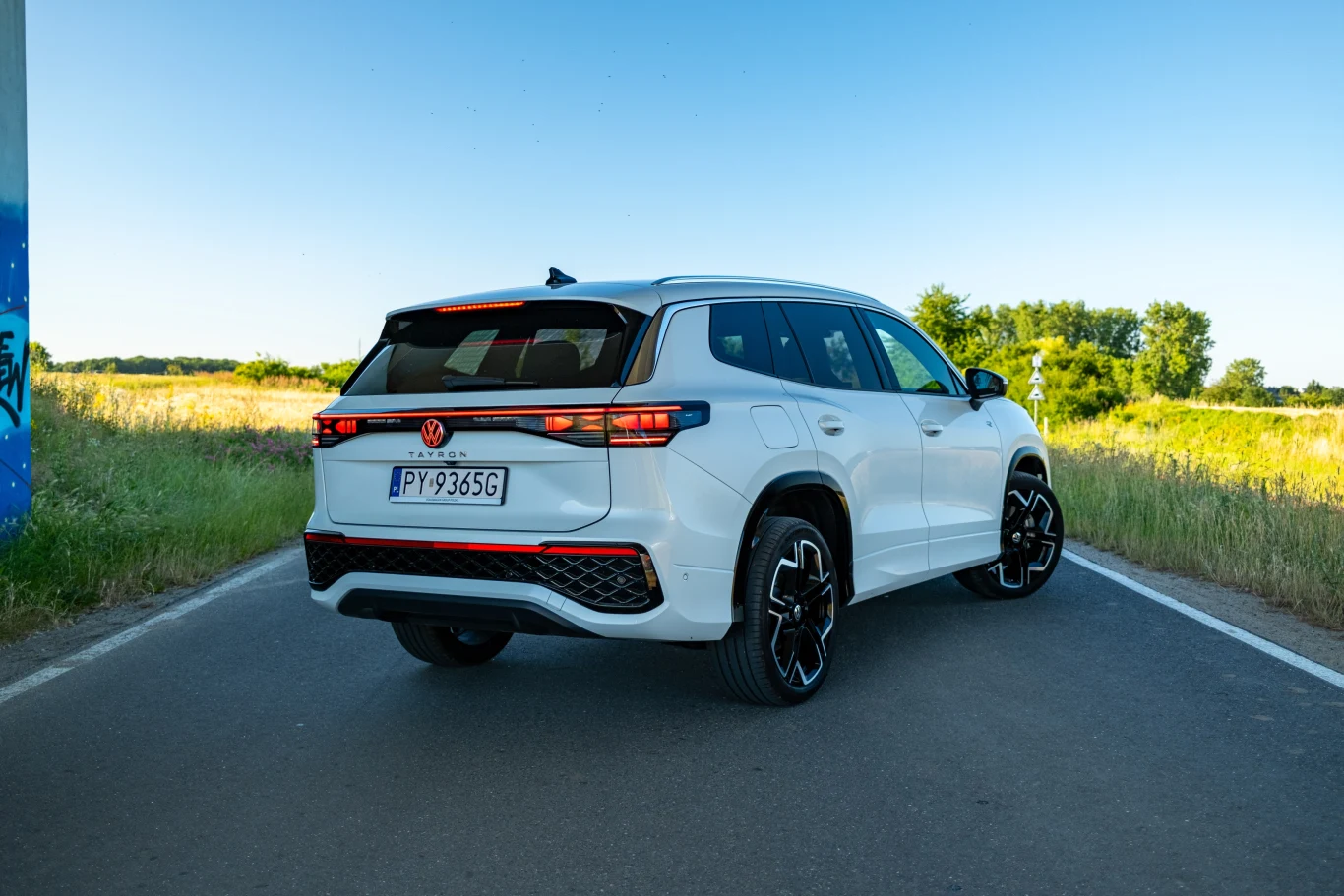 Tył auta wyróżnia się podświetlanym na czerwono logo Volkswagena. Nowoczesny, biały SUV zaparkowany na wiejskiej drodze otoczonej zielenią, widoczne eleganckie felgi i sportowe akcenty, w tle pola i drzewa przy jasnym, bezchmurnym niebie.