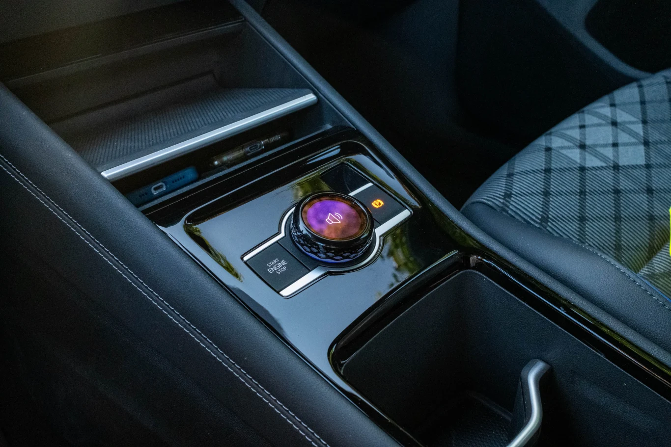 Pokrętło do regulacji głośności jest pod ręką, po wciśnięciu można wybierać nim tryb jazdy lub “atmosferę” wnętrza, czyli między innymi rodzaj nastrojowego oświetlenia. Nowoczesna konsola środkowa samochodu z elektroniczną gałką zmiany biegów oraz detalami wykończenia, częściowo widoczne siedzenie pokryte kratkowanym materiałem.