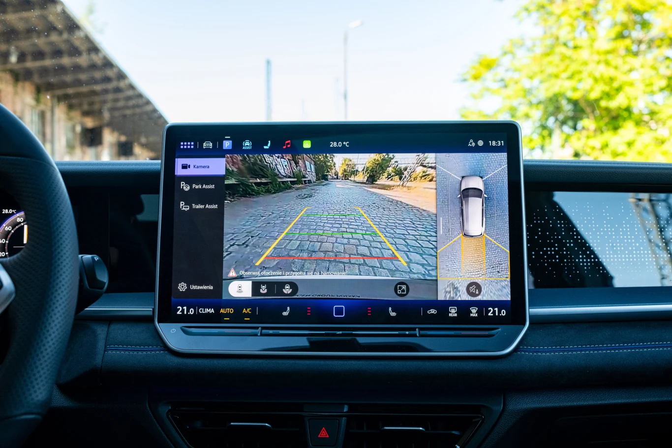 Obraz z tylnej kamery cofania powinien być lepszej jakości, na dużym ekranie wyraźnie widać “zaśnieżony” obraz. Ekran systemu multimedialnego samochodu wyświetla obraz z kamery cofania wraz z wyznaczonymi liniami pomocniczymi i widokiem z góry na pojazd, w tle fragment wnętrza auta i elementy otoczenia drogi.