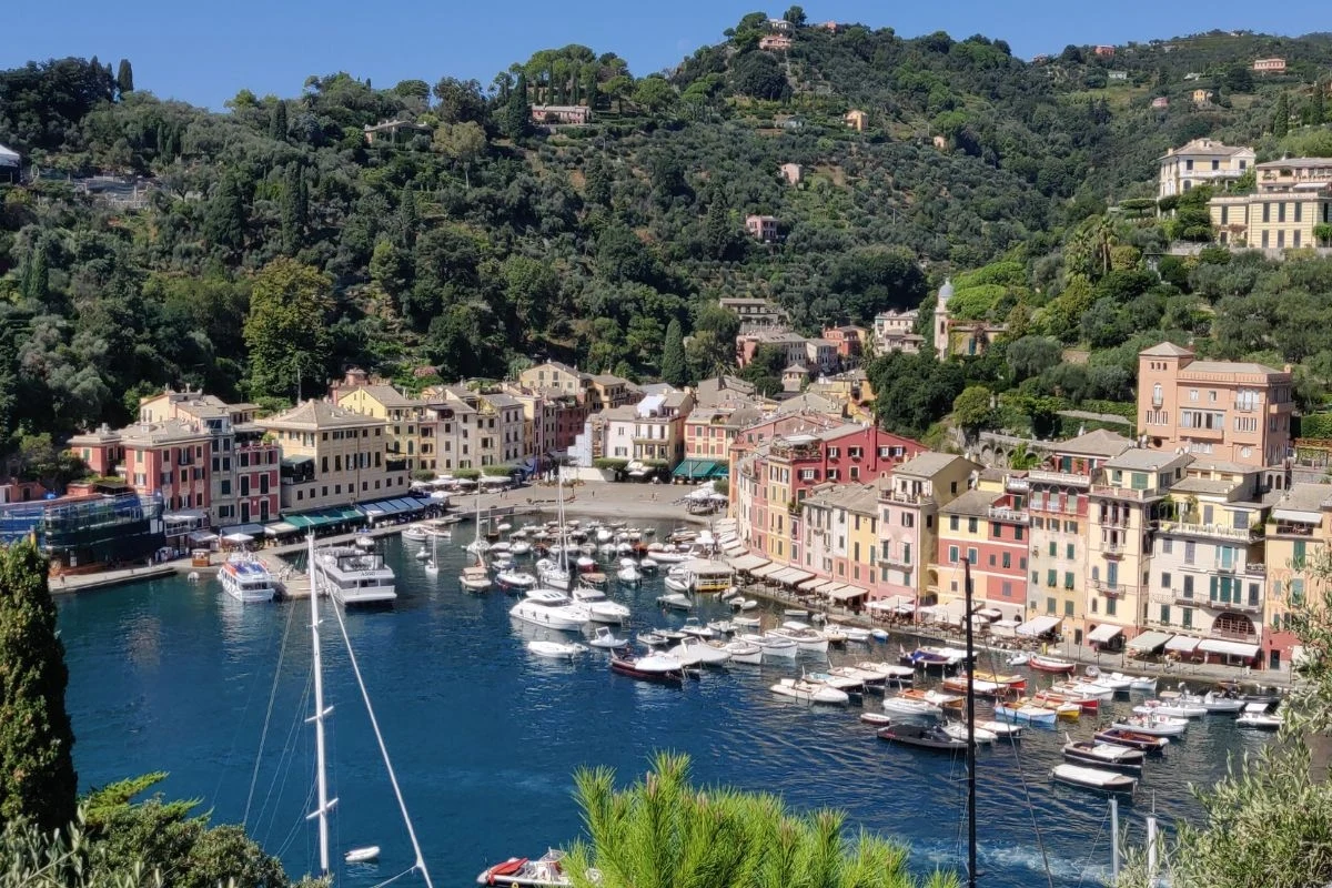 Kolorowe kamienice ułożone przy porcie z luksusowymi jachtami i łodziami, otoczone zielonymi wzgórzami, całość tworzy malowniczy śródziemnomorski krajobraz.