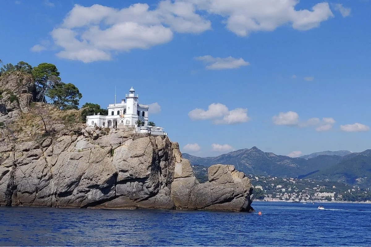 Biały budynek latarni morskiej stojący na wysokim skalistym klifie nad błękitnym morzem, w tle łagodne, zielone wzgórza oraz fragment zabudowań miejskich, nad wszystkim błękitne niebo z pojedynczymi chmurami.