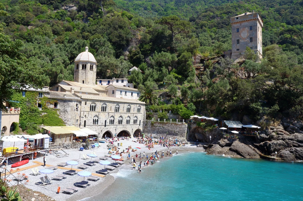 San Fruttuoso w pełnej okazałości Plaża z turkusową wodą, turyści kąpiący się i wypoczywający na leżakach, zabytkowy klasztor z kopułą i arkadami przy plaży, z tyłu wysoka wieża otoczona bujną zielenią.