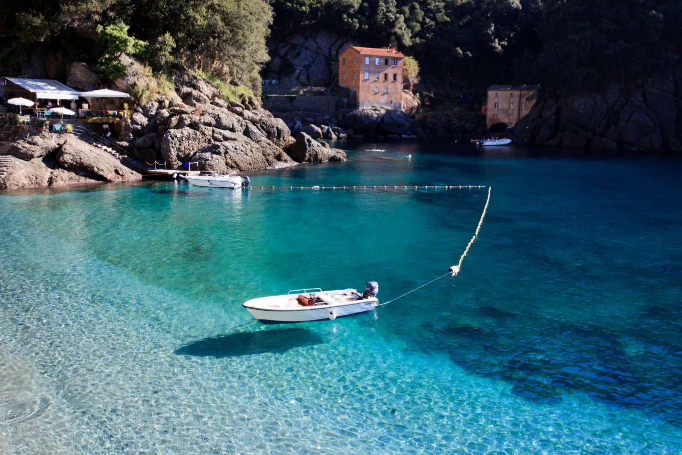 Mała łódź zacumowana na przejrzystej, turkusowej wodzie zatoki, w tle skaliste wybrzeże porośnięte drzewami oraz dwa jasne budynki położone na brzegu.