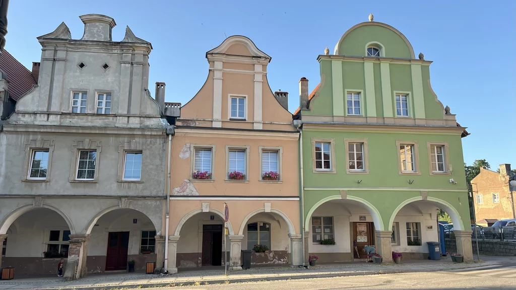 Rynek w Chełmsku Śląskim otaczają zabytkowe barokowe kamienice z podcieniami Rząd trzech zabytkowych kamienic o pastelowych fasadach z arkadami na parterze, ozdobione detale architektoniczne oraz okna z kwiatami, położone w słonecznym mieście.