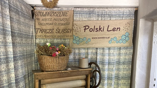 Stare drewniane narzędzie do obróbki lnu ustawione przed zasłonami, na nim wiklinowy kosz z suchymi kwiatami oraz dekoracyjny motek przędzy. W tle dwa bannery, jeden informacyjny a drugi promocyjny z napisem Polski Len.