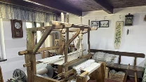 Drewniane krosno do tkania ustawione w tradycyjnym, wiejskim wnętrzu z prostymi meblami, ławką, krzesłem z kolorową poduszką oraz dekoracjami na ścianach, obok skrzynia z przędzą, całość utrzymana w rustykalnym stylu.