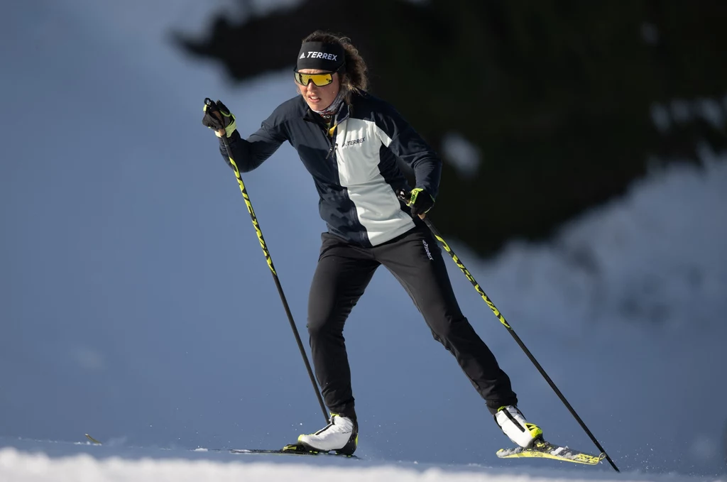 Laura Dahlmeier Sportowiec w ciemnym stroju zimowym i okularach przeciwsłonecznych biegnie na nartach biegowych pośnieżonej trasie w słoneczny dzień, używając kijków narciarskich.