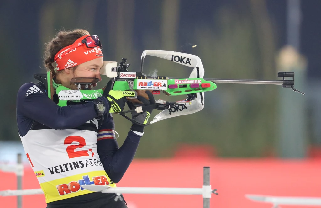 Laura Dahlmeier Zawodniczka biathlonu w sportowym stroju, z numerem startowym i opaską na głowie, celuje z karabinu podczas zawodów, w tle rozmyte elementy stadionu.
