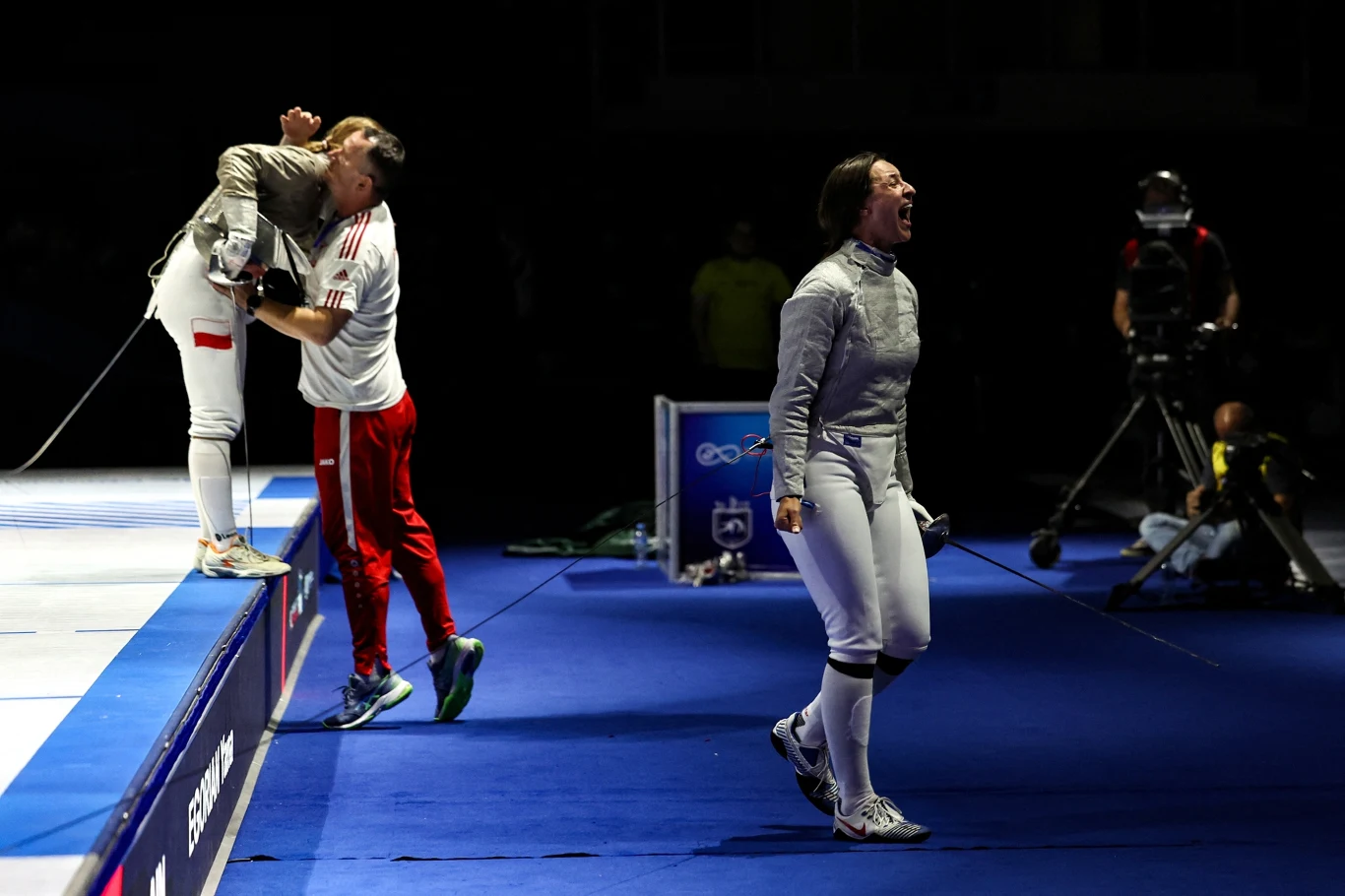 Zuzanna Cieślar zdobyła srebrny medal MŚ w szermierce. W finale uległa Rosjance Silnie oświetlona scena sportowa podczas zawodów szermierki, fencerka w białym stroju wyraża ogromne emocje po zakończonej walce, w tle drugi zawodnik i trener okazujący radość na podwyższeniu, atmosfera podkreślona dynamicznym światłem i ciemnym tłem.