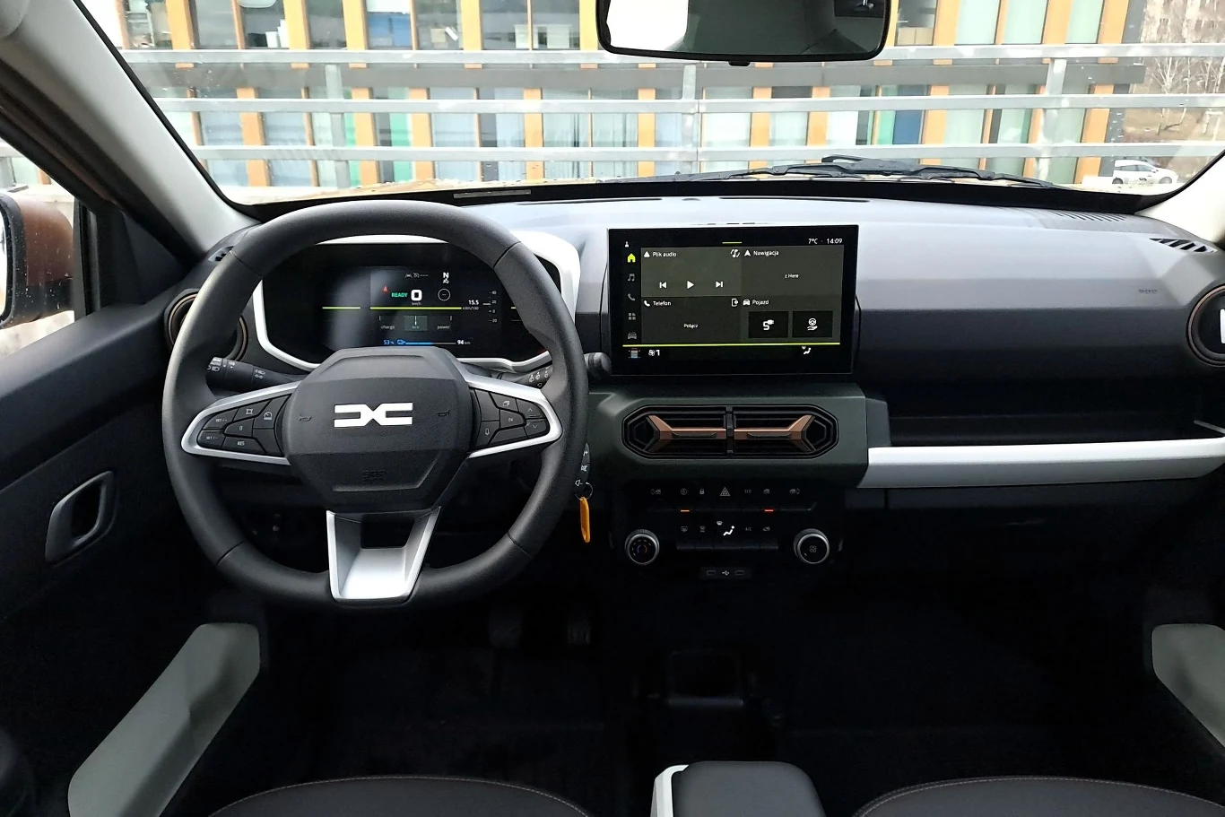 Kokpit nowoczesnego samochodu z cyfrowymi wyświetlaczami, wielofunkcyjną kierownicą z logo DC i dużym ekranem dotykowym centralnym, wnętrze utrzymane w minimalistycznej stylistyce z prostymi przyciskami do sterowania klimatyzacją i systemem multimedial...