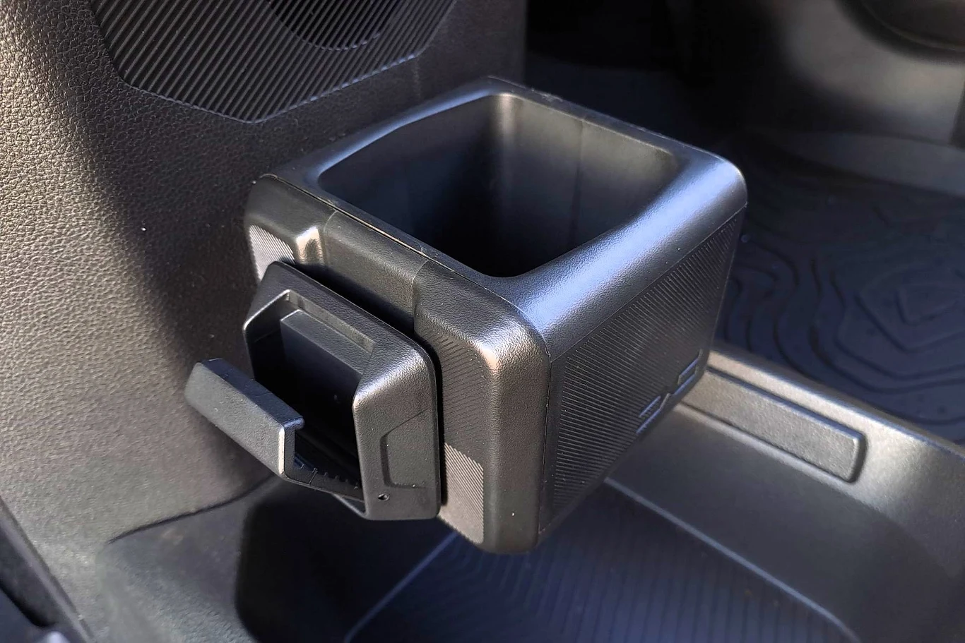 Czarny plastikowy uchwyt na napoje zamocowany przy bocznej ścianie wnętrza samochodu, widoczne także fragmenty tapicerki i podłogi z gumową matą.