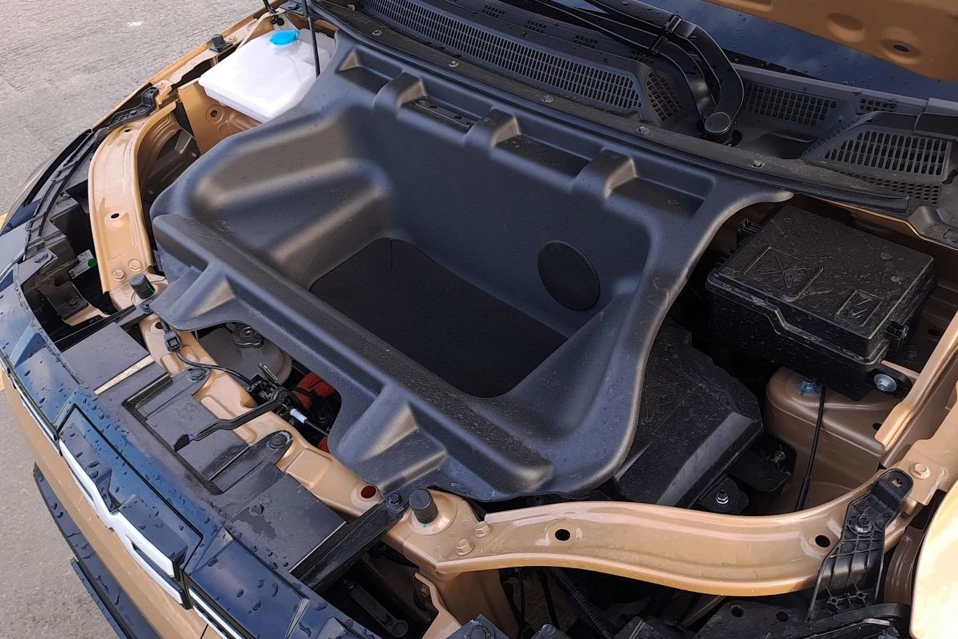 Komora bagażowa z przodu samochodu elektrycznego, widoczny czarny plastikowy kufer oraz elementy techniczne pod maską, zlewka płynu chłodniczego z niebieską nakrętką po lewej stronie.
