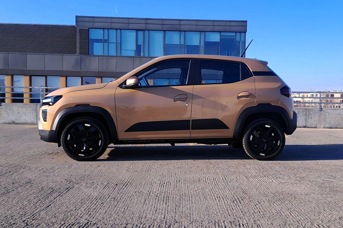 Dacia Spring Brązowy kompaktowy SUV stoi na parkingu z betonową nawierzchnią na tle nowoczesnego, przeszklonego budynku.