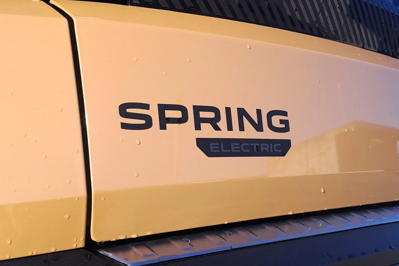 Logo SPRING ELECTRIC umieszczone na żółtej karoserii samochodu osobowego, widoczne detale lakieru oraz kropelki wody po myciu auta.
