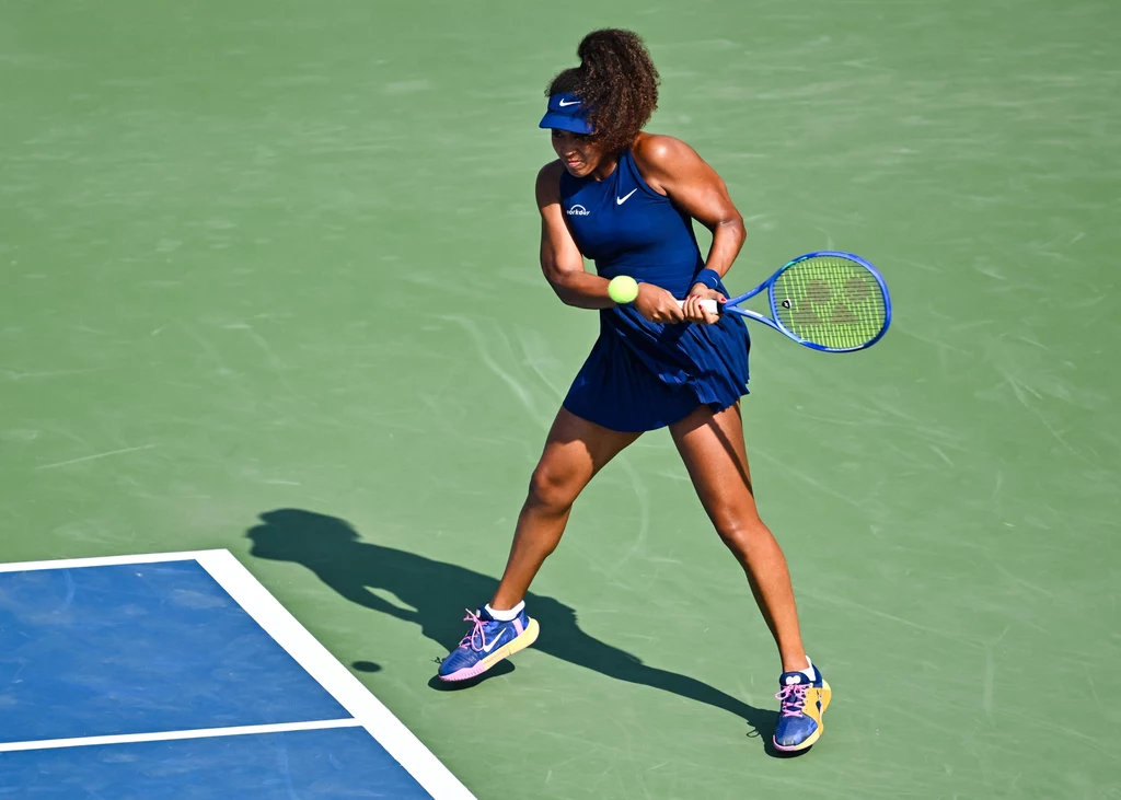 Naomi Osaka Tenisistka ubrana w granatowy strój sportowy wykonuje dynamiczne uderzenie rakietą tenisową, piłka znajduje się tuż przed nią, a całość odbywa się na zielonym korcie tenisowym.