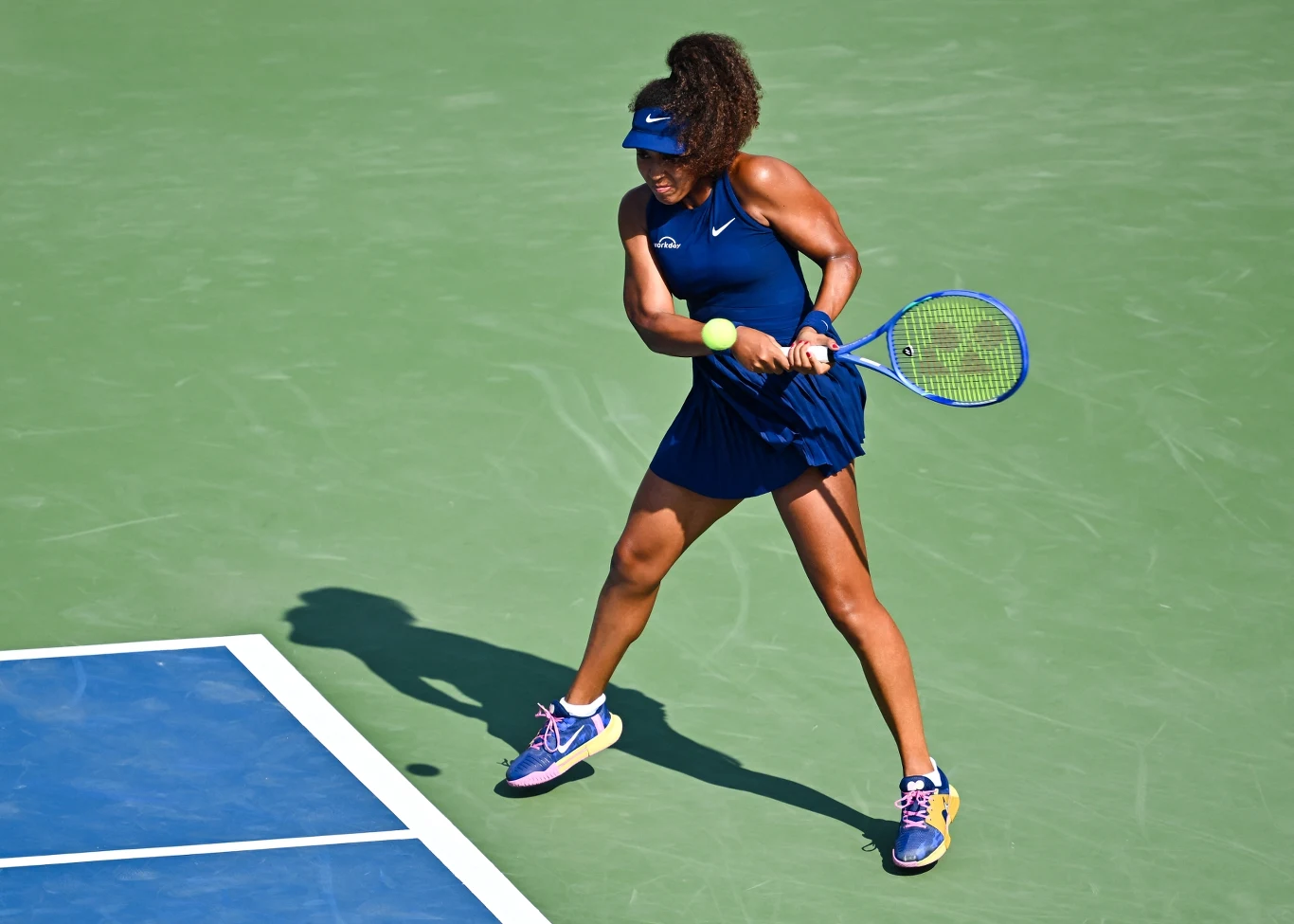 Tenisistka ubrana w granatowy strój sportowy wykonuje dynamiczne uderzenie rakietą tenisową, piłka znajduje się tuż przed nią, a całość odbywa się na zielonym korcie tenisowym.