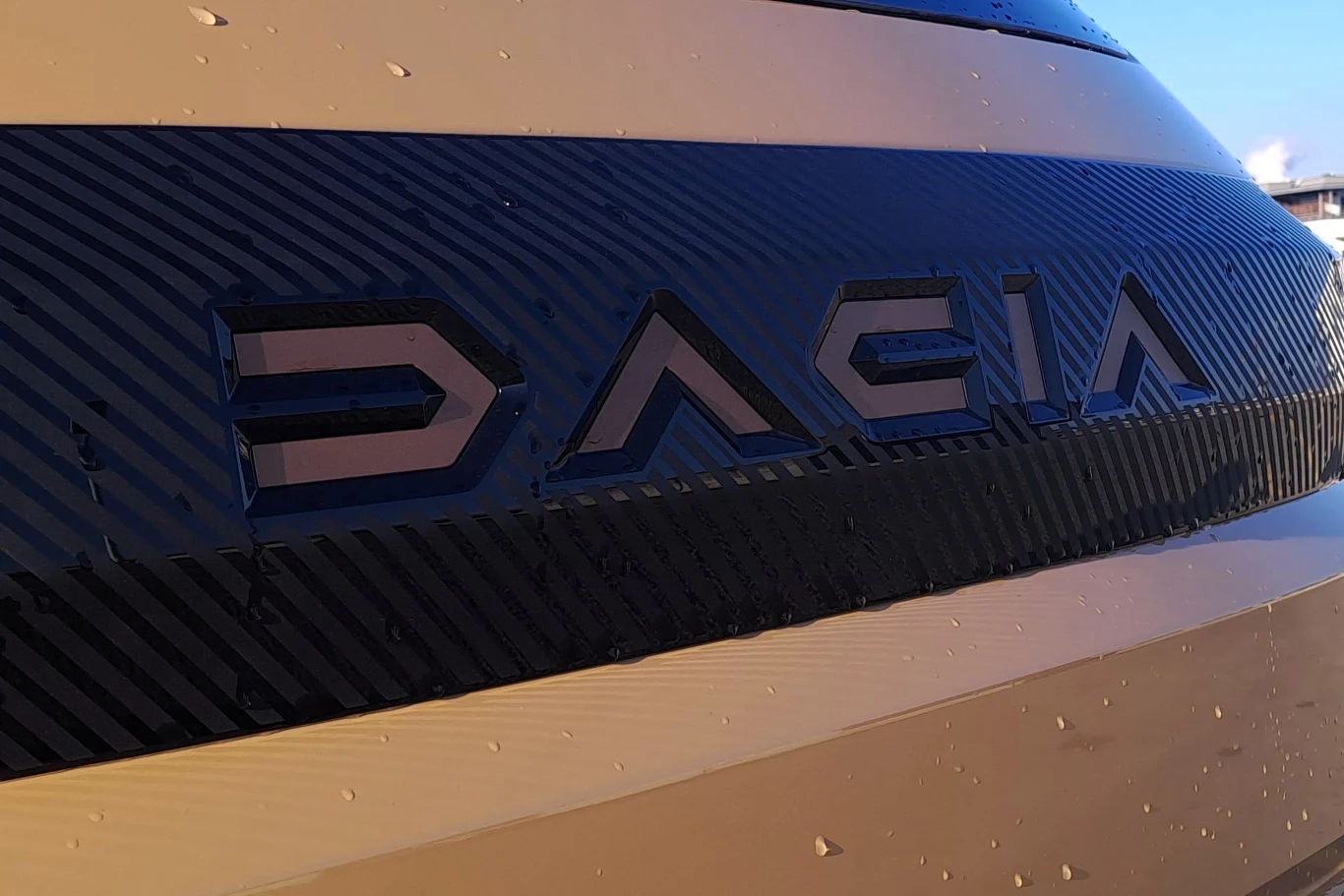 Logo marki Dacia umieszczone na tylnej klapie samochodu, otoczone wyraźnymi elementami stylistycznymi oraz drobnymi kroplami wody.