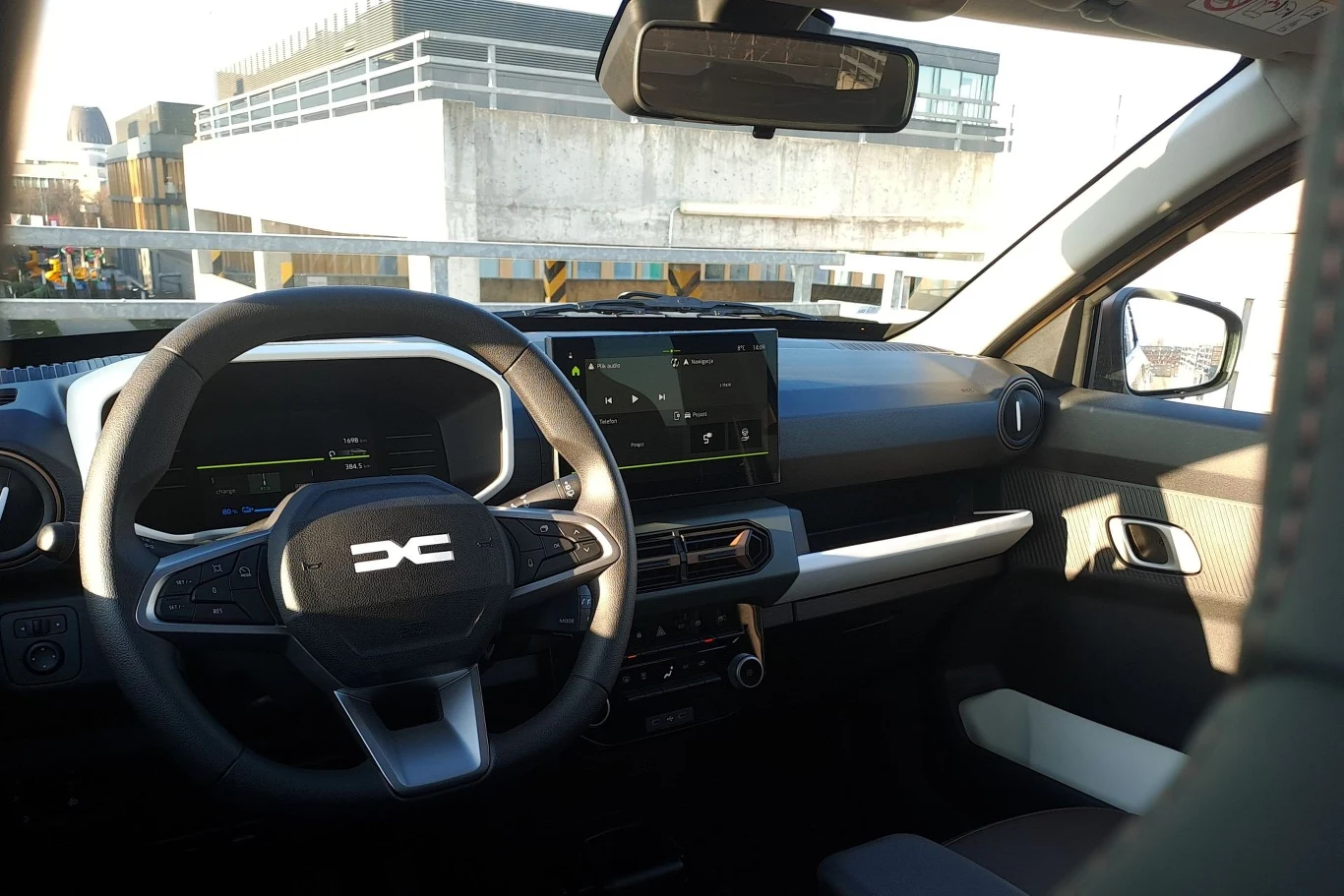 Nowoczesna deska rozdzielcza samochodu z dużym wyświetlaczem na środku, cyfrowymi wskaźnikami, kierownicą z logo marki oraz jasnymi tapicerkami siedzeń. Za szybą widać część infrastruktury miejskiej.