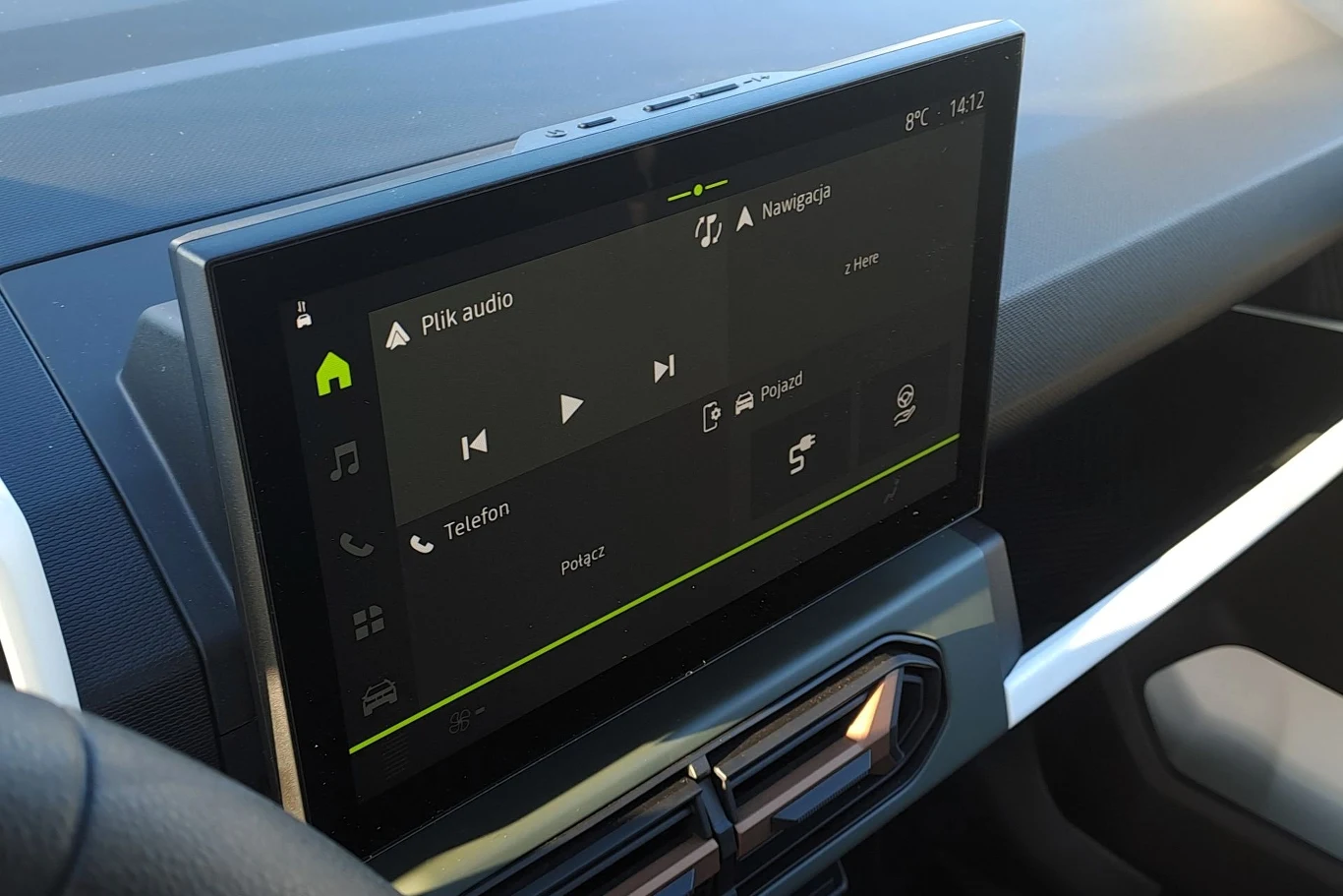 Dotykowy ekran systemu multimedialnego samochodu z wyświetlonymi opcjami audio, nawigacji i telefonu, utrzymany w nowoczesnym stylu, zamontowany na desce rozdzielczej obok kierownicy.