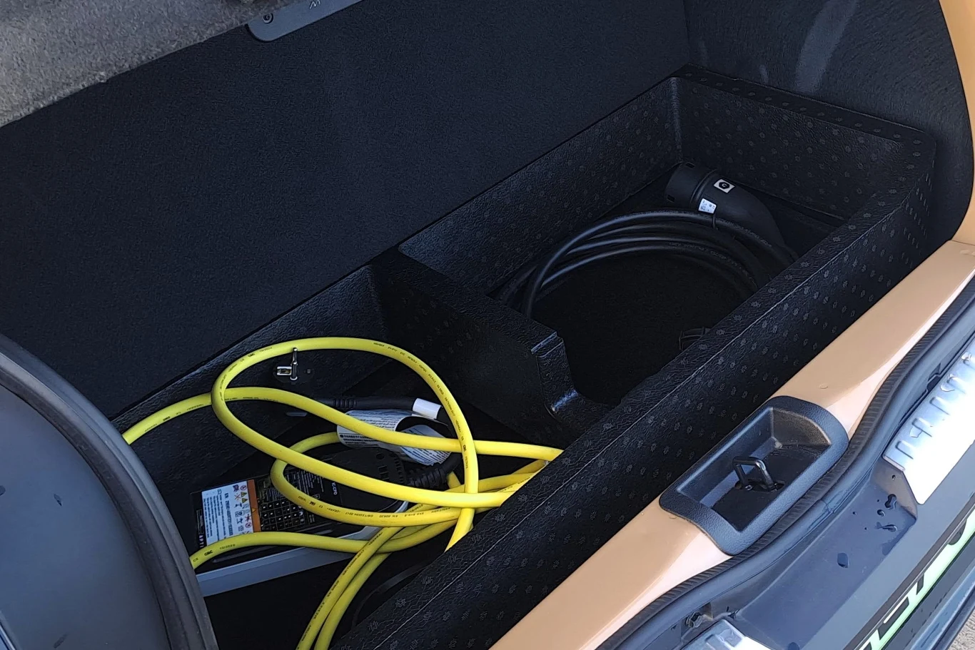 Schowek w bagażniku samochodu z widocznymi przewodami do ładowania pojazdu elektrycznego, ułożonymi w przegródkach i zabezpieczonymi w czarnym, wyściełanym wnętrzu.