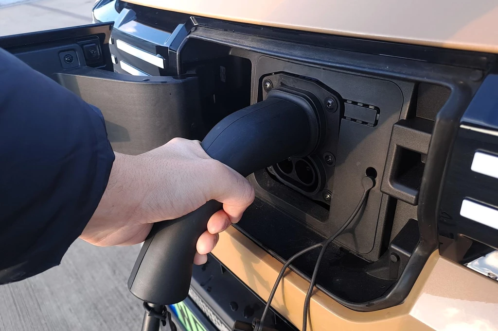 Ręka trzymająca wtyczkę i podłączająca ją do portu ładowania samochodu elektrycznego, widoczny przedni panel pojazdu i szczegóły technologiczne związane z procesem ładowania.