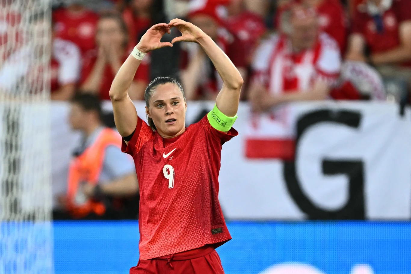 Piłkarka w czerwonym stroju z opaską kapitańską unosi ręce nad głową, tworząc gest serca, w tle kibice ubrani w czerwono-białe stroje oraz fragment bannera stadionowego.