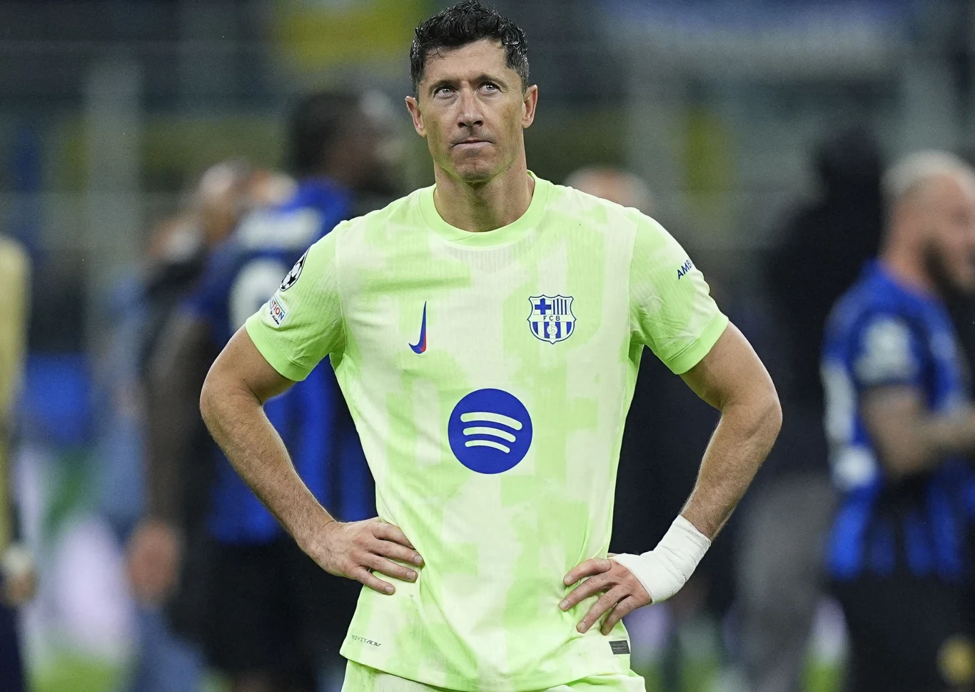 Piłkarz w jasnozielonej koszulce z logo hiszpańskiego klubu FC Barcelona oraz sponsorem na klatce piersiowej, stojący na boisku z rękami opartymi na biodrach, w tle inni zawodnicy rozmazani.