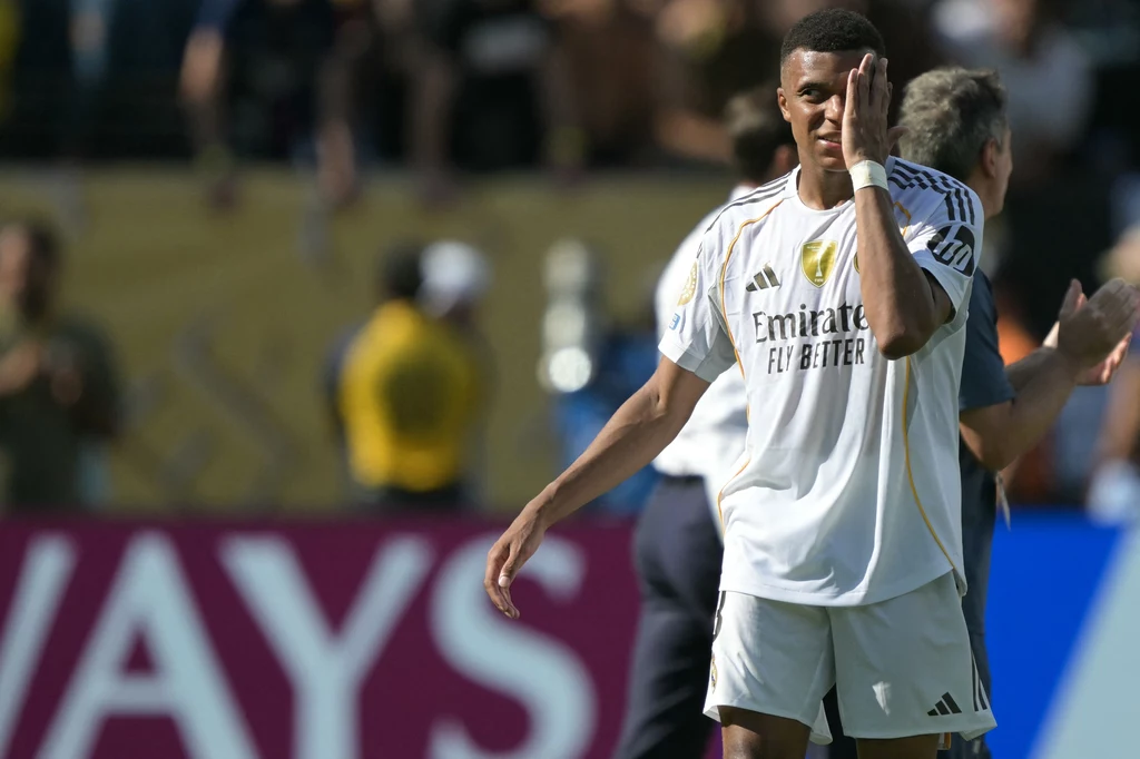 Kylian Mbappe Piłkarz w białej koszulce z logo sponsora dotyka twarzy podczas meczu piłkarskiego, w tle rozmazane sylwetki innych osób i stadion.