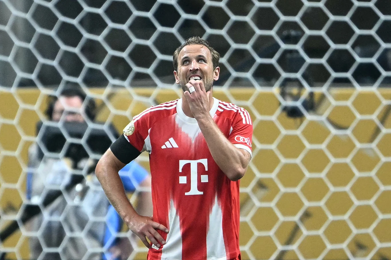 Zawodnik drużyny piłkarskiej w czerwono-białej koszulce z logotypem sponsora stoi na tle siatki bramki, z wyrazem zamyślenia na twarzy, oparty o biodro, w tle rozmazane postacie i stadion.