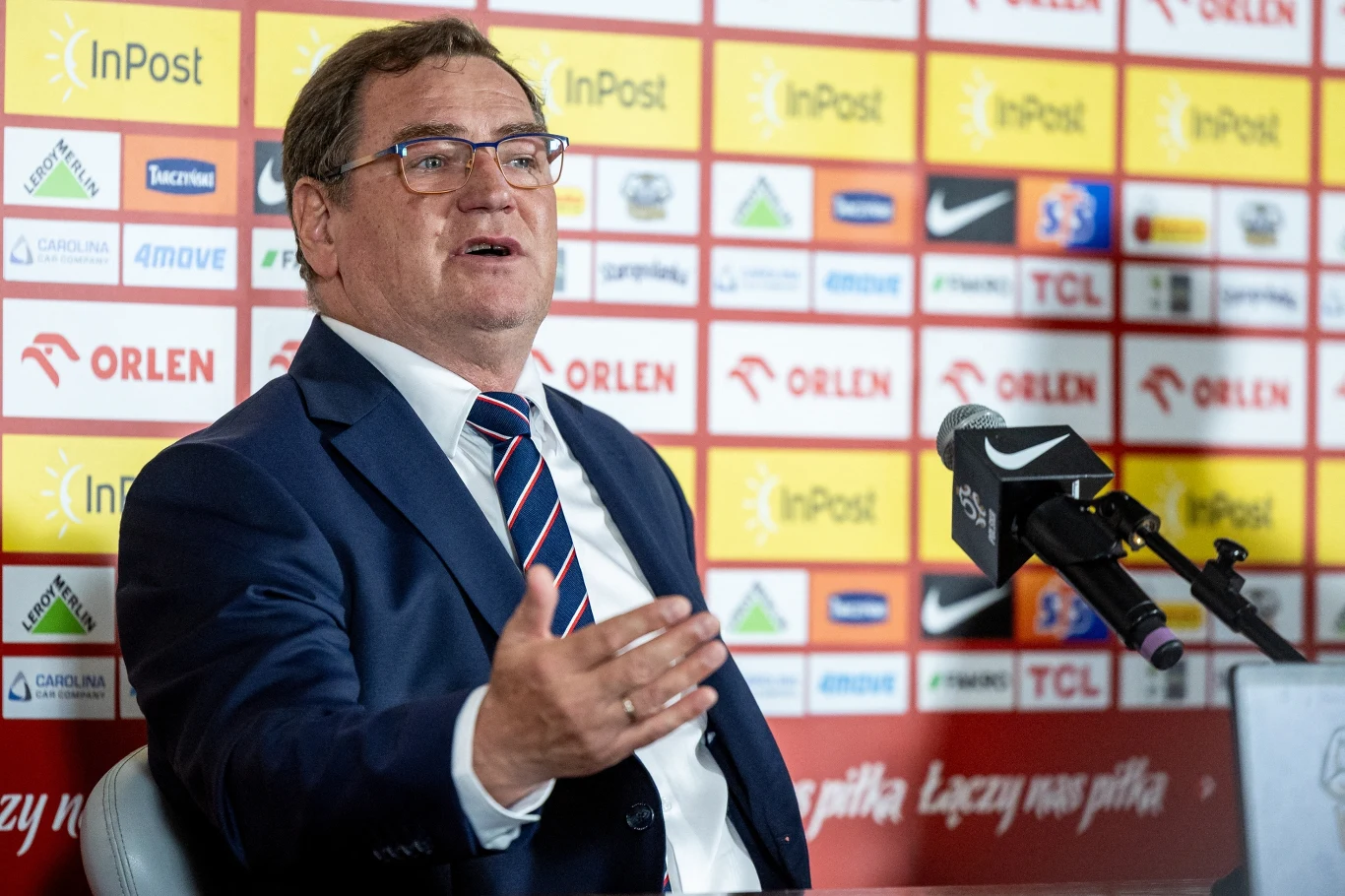 Mężczyzna w garniturze rozmawiający podczas konferencji prasowej, z mikrofonem przed sobą, na tle ściany z logotypami sponsorów