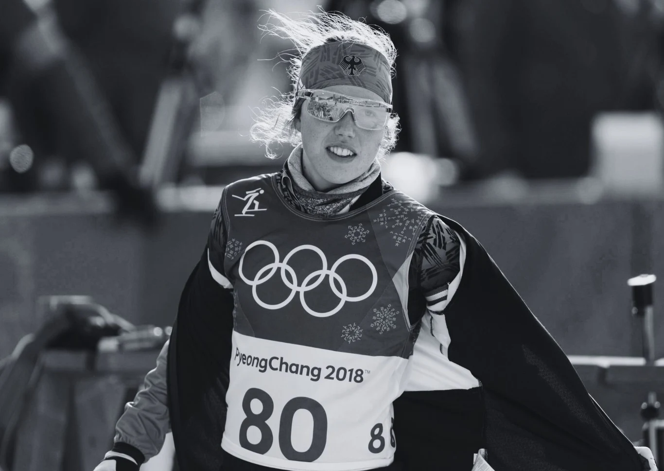 Laura Dahlmeier Sportowiec ubrany w strój narciarski z numerem 80 i charakterystyczną kamizelką z logo igrzysk olimpijskich prezentuje się na trasie olimpijskiej zimowych igrzysk w Pjongczangu 2018, z wyraźnie widocznym medalionem i śniegowym motywem wokół.