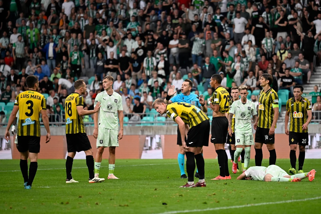 Kajrat Ałmaty pierwszy raz w swojej historii awansował do trzeciej rundy eliminacji Ligi Mistrzów Kilku piłkarzy w żółto-czarnych oraz jasnozielonych strojach zgromadzonych na boisku, jeden z zawodników leży na murawie, wokół niego toczy się dyskusja z udziałem sędziego i reszty zawodników, trybuny wypełnione kibicami.