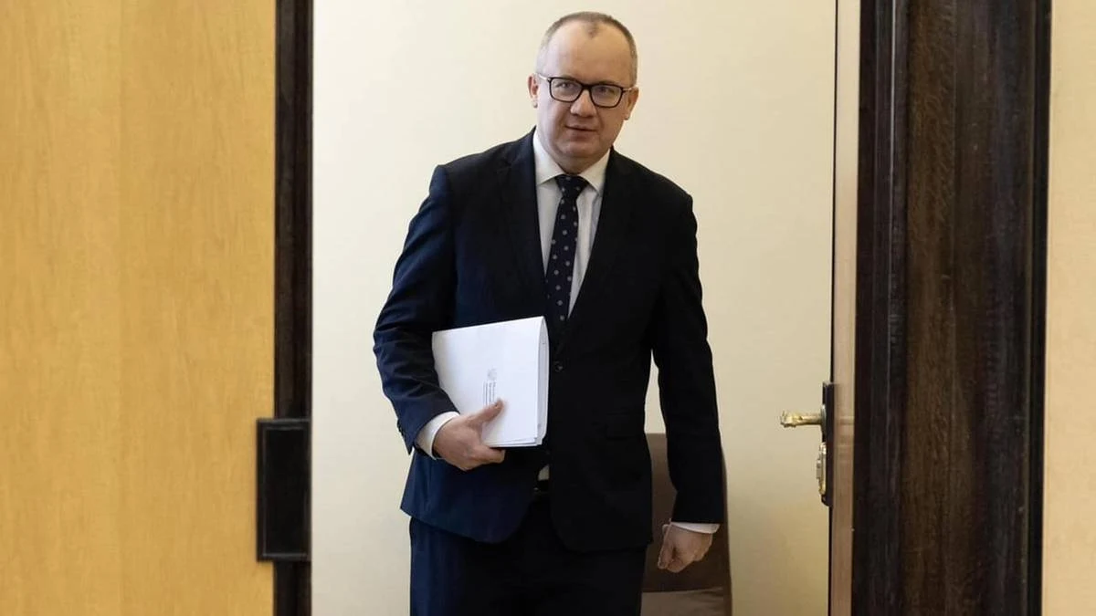 Adam Bodnar Mężczyzna w garniturze z teczką w dłoni wchodzi do pomieszczenia przez otwarte drzwi, neutralne wnętrze, elegancki ubiór, oficjalny charakter sytuacji.