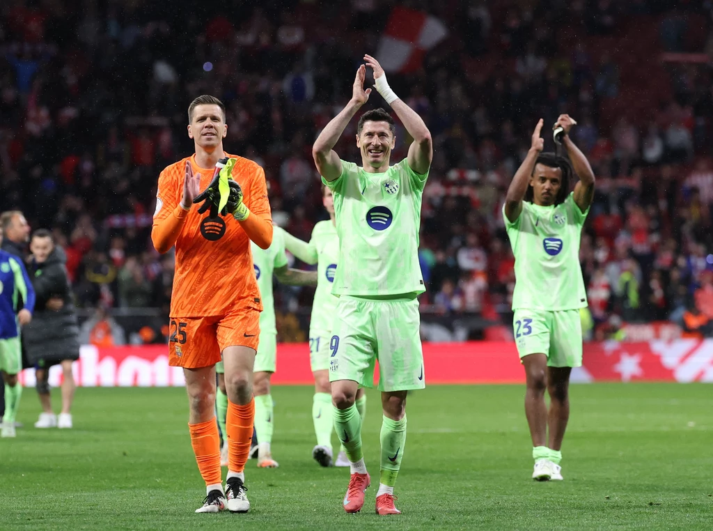 Wojciech Szczęsny, Robert Lewandowski i Jules Kounde w barwach FC Barcelona Trzech piłkarzy w jasnych strojach sportowych oraz bramkarz w pomarańczowym stroju opuszczają boisko, bijąc brawo publiczności, za nimi widać rozmazany tłum kibiców i stadionowe światła.
