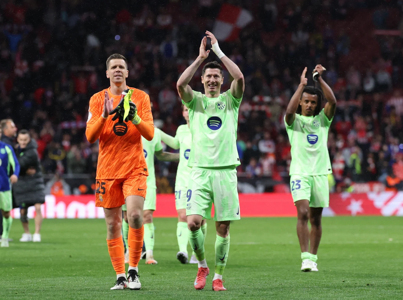 Wojciech Szczęsny, Robert Lewandowski i Jules Kounde w barwach FC Barcelona Trzech piłkarzy w jasnych strojach sportowych oraz bramkarz w pomarańczowym stroju opuszczają boisko, bijąc brawo publiczności, za nimi widać rozmazany tłum kibiców i stadionowe światła.