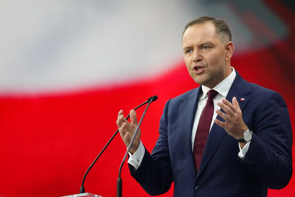 Karol Nawrocki Mężczyzna w garniturze przemawia zza mównicy gestykulując dłońmi na tle rozmytej biało-czerwonej flagi Polski.