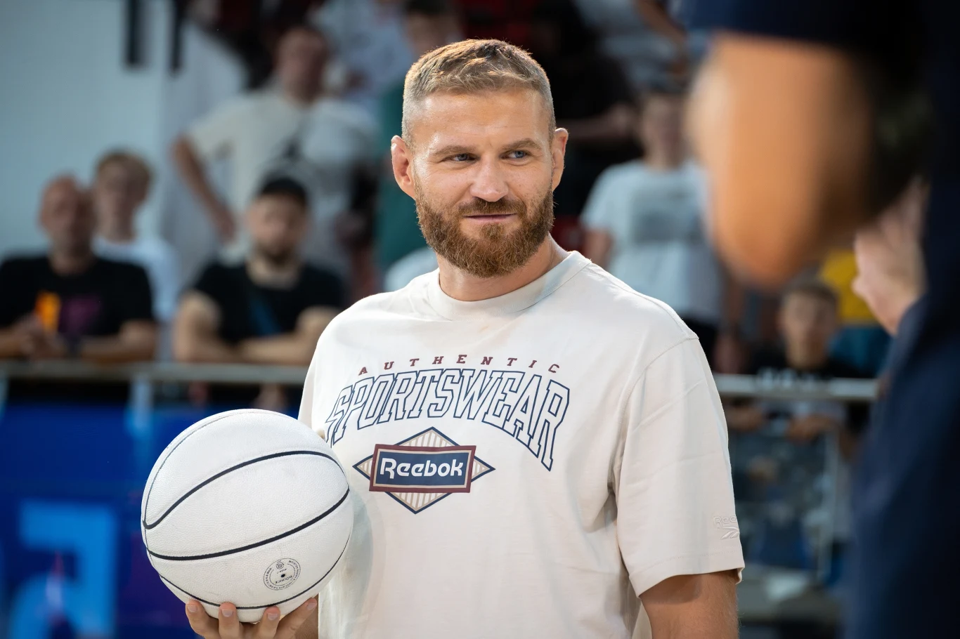 Jan Błachowicz Mężczyzna z brodą w jasnej koszulce sportowej marki Reebok trzyma biały koszykarski piłkę, stojąc na hali sportowej wśród rozmazanego tłumu kibiców na trybunach.
