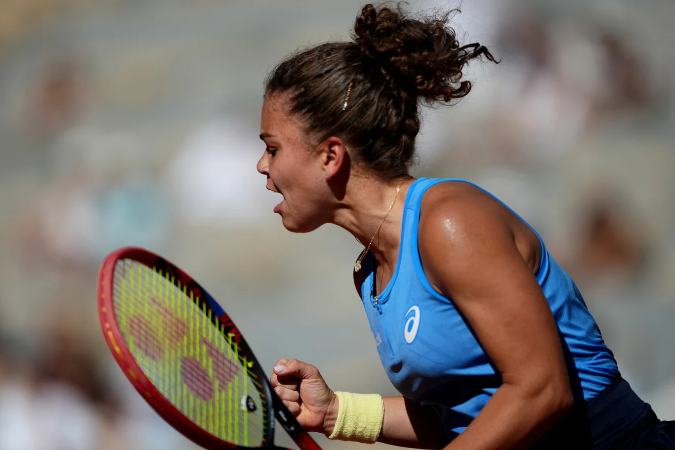 Zawodniczka ubrana w niebieski sportowy top z włosami spiętymi w koka energicznie ściska dłoń na korcie tenisowym, okazując silne emocje po rozegranym punkcie, w tle rozmyte sylwetki widzów.
